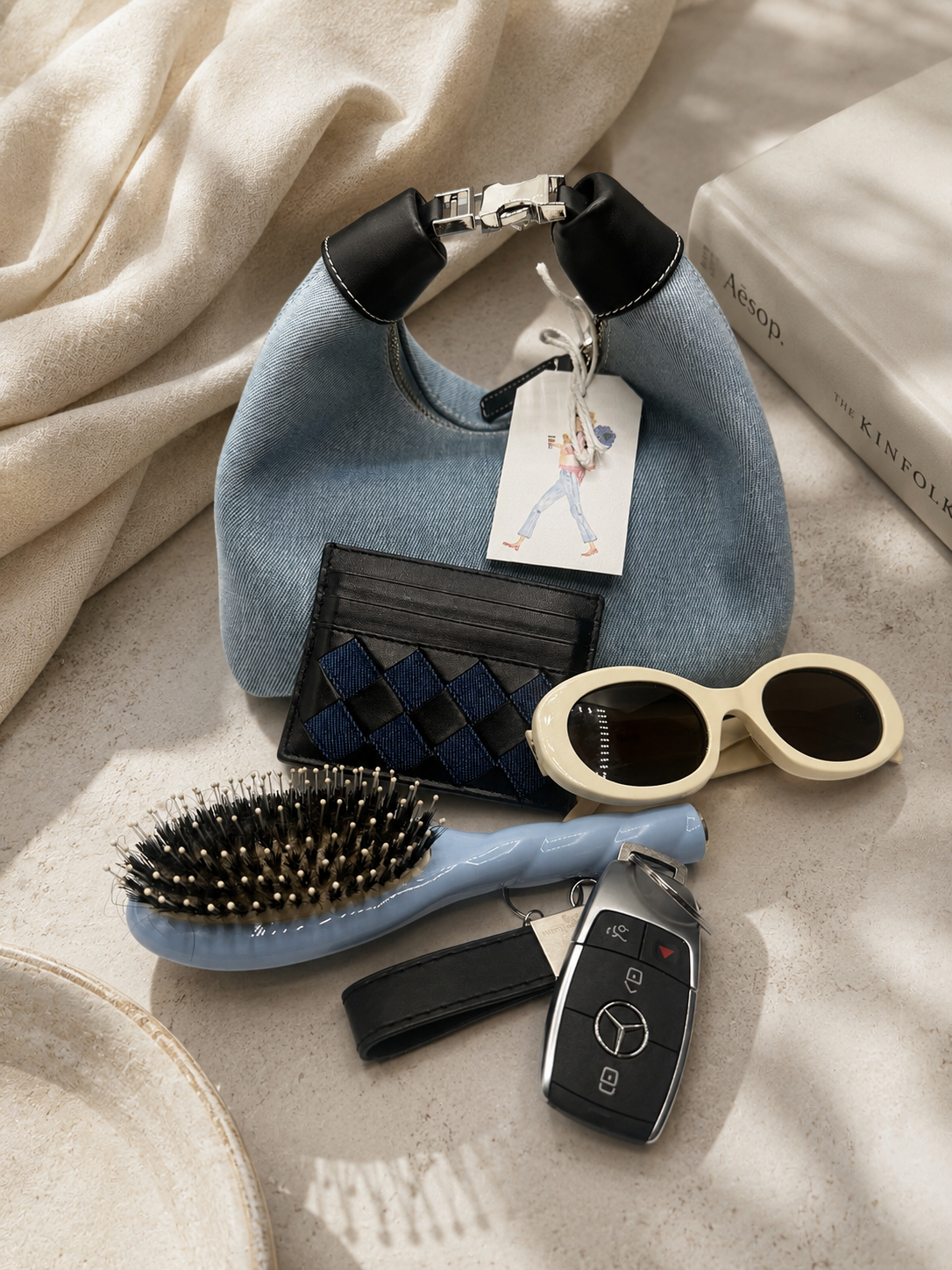 A styled flat-lay featuring a light blue denim shoulder bag with black leather trim and a hanging tag, surrounded by accessories including cream sunglasses, a blue hairbrush, a black quilted card holder, a Mercedes key fob, and draped cream fabric with a book in the background.
